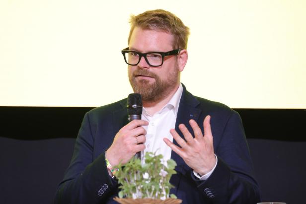 Ein Mann mit Bart und Brille spricht in ein Mikrofon und gestikuliert, vor ihm steht ein Topf mit grünen Pflanzen.