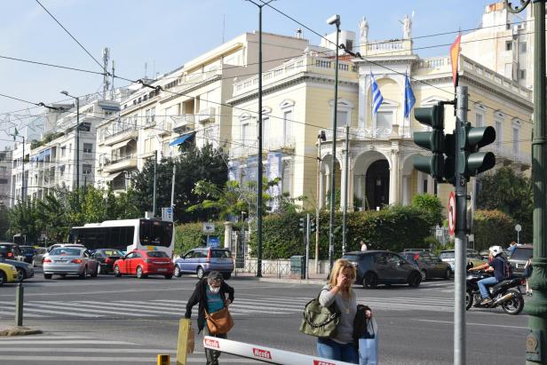 Kreuzung in Athen mit Autos, Bus, Fußgängern und Ampeln vor historischen Gebäuden.