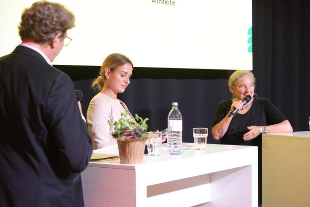 Drei Personen diskutieren an einem Tisch mit Mikrofonen, Wasserflaschen und einem Blumentopf vor dunklem Hintergrund.