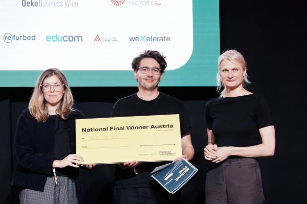 Drei Personen stehen nebeneinander, eine hält einen großen gelben Scheck mit der Aufschrift „National Final Winner Austria“.