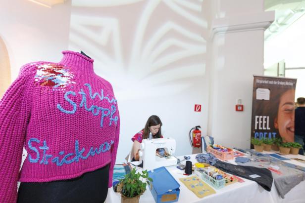 Eine Frau näht an einer Nähmaschine an einem Tisch mit Nähutensilien, im Vordergrund steht eine pinke Strickjacke auf einer Schneiderpuppe.