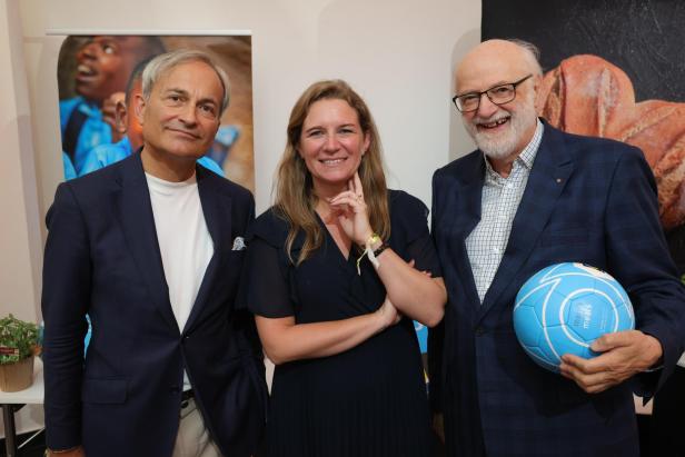 Zwei Männer und eine Frau posieren vor Plakaten, wobei einer der Männer einen blauen Fußball der Organisation Mary’s Meals hält.