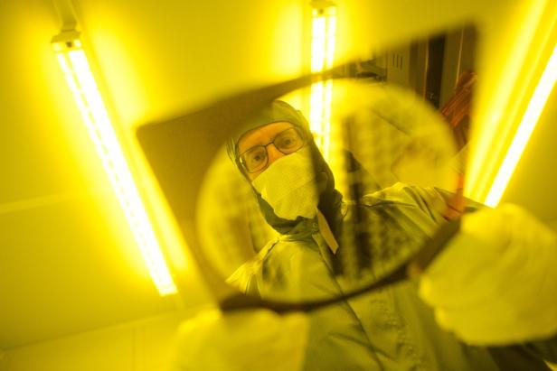 Person in Schutzkleidung betrachtet eine runde Scheibe unter gelbem Licht in einem Reinraum.