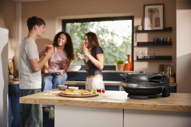 Kenwood Pizzaofen und Pizza im Vordergrund - dahinter 2 Frauen und 1 Mann beim Pizzaessen und plaudern 