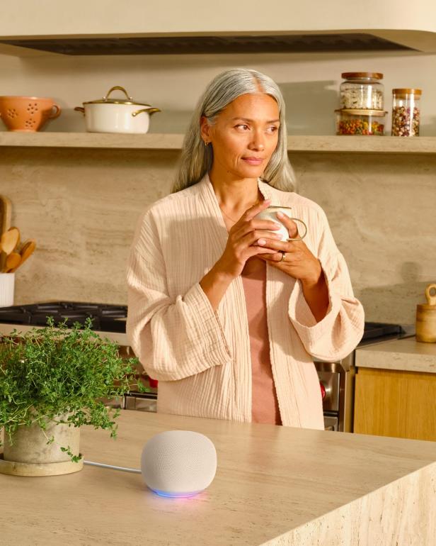 Eine Frau steht in einer Küche neben dem neuen Google Home Speaker mit leuchtendem Ring.