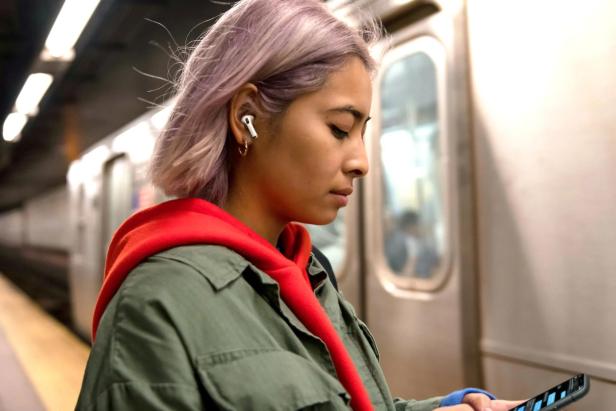 Eine junge Frau mit lila Haaren und Kopfhörern schaut in einer U-Bahn-Station auf ihr Smartphone.