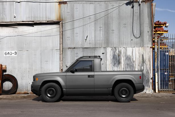 Ein grauer, kantiger Slate-E-Pick-up mit kurzer Ladefläche steht vor einer Wellblechwand.