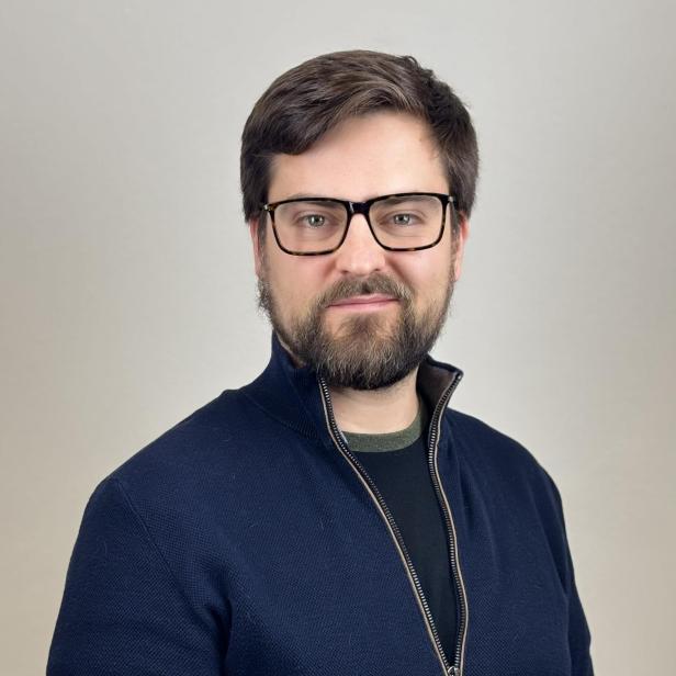 Mann mit Bart und Brille in dunkelblauer Jacke vor hellem Hintergrund.