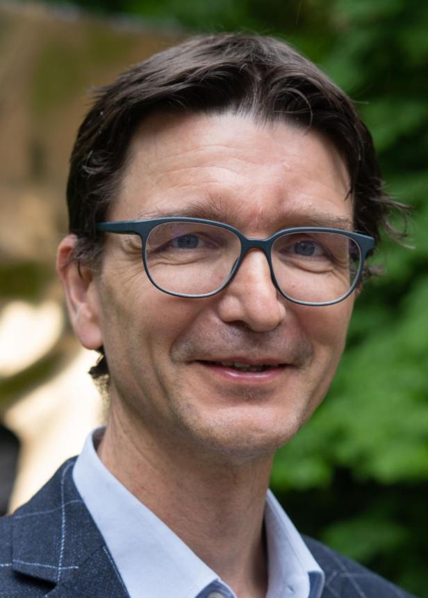 Nahaufnahme eines lächelnden Mannes mit Brille im Anzug vor grünem Hintergrund.