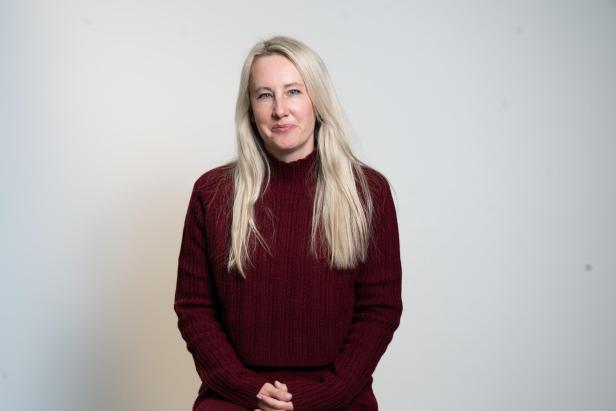 Eine blonde Frau im bordeauxfarbenen Pullover sitzt vor hellem Hintergrund und blickt in die Kamera.