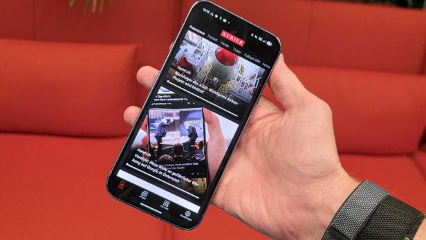 Hand hält ein Smartphone mit KURIER-News-App vor rotem Sofa.