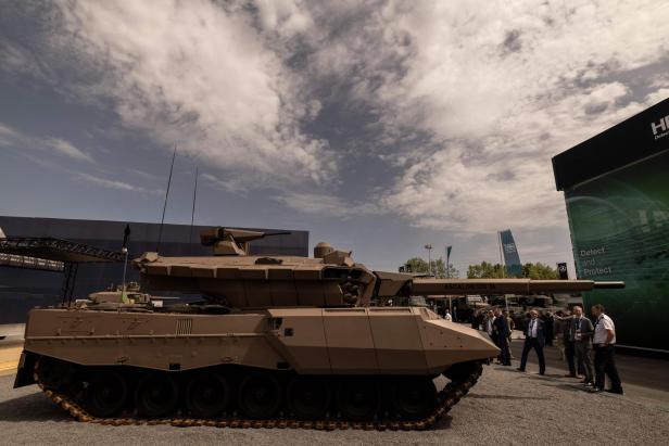 Ein modernes Kampffahrzeug mit langem Geschütz steht auf einer Messe, daneben stehen mehrere Anzugträger.