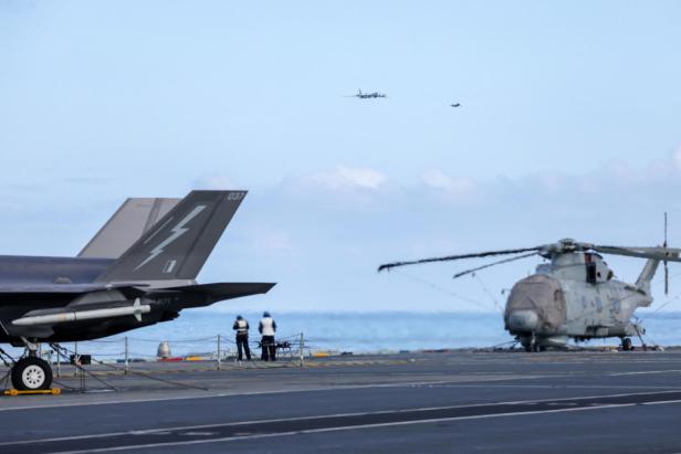 Flugdeck eines Flugzeugträgers mit Jets, einem Hubschrauber und zwei Personen, darüber zwei Flugzeuge am Himmel.