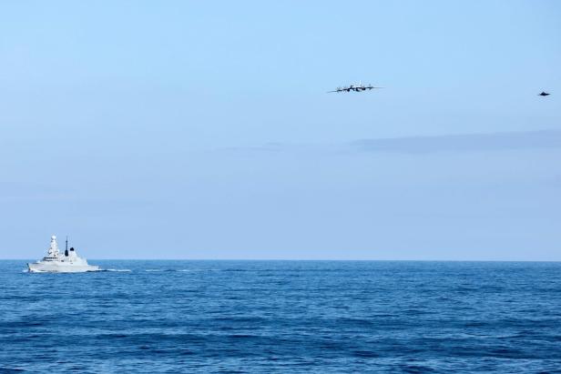 Ein Kriegsschiff fährt auf dem Meer, während ein Propellerflugzeug und ein Kampfjet am blauen Himmel darüber fliegen.
