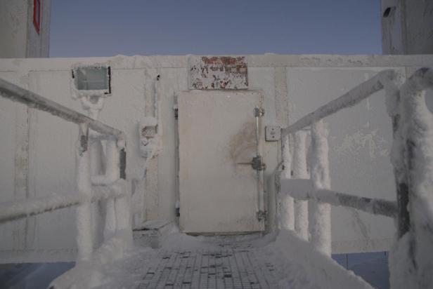 Eine von Eis und Schnee bedeckte Tür führt in ein Gebäude, flankiert von einem vereisten Geländer unter einem klaren Himmel.
