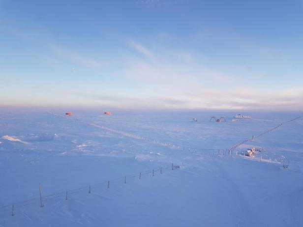Weite, schneebedeckte Eislandschaft der Antarktis mit kleinen Forschungsstationen und einem Zaun unter einem hellblauen Himmel.
