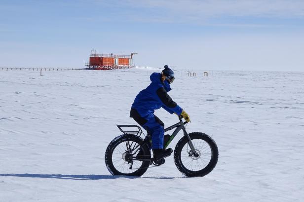 Eine Person in blauer Winterkleidung fährt mit einem Fatbike durch eine weite, schneebedeckte Landschaft vor einer orangefarbenen Forschungsstation.