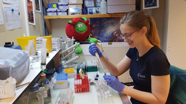 Eine lächelnde ESA-Mitarbeiterin arbeitet mit einer Pipette an einem Labortisch, auf dem sich zahlreiche Reagenzgläser und ein Stofftier befinden.