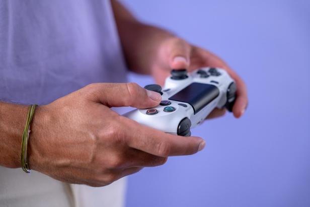 Eine Person hält einen weißen PlayStation-Controller vor einem hellvioletten Hintergrund in den Händen.