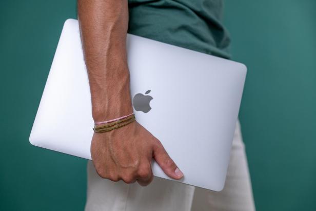 Eine Person trägt ein silbernes Apple MacBook unter dem Arm vor einem grünen Hintergrund.
