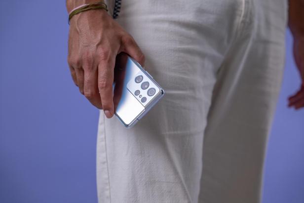 Eine Person in heller Hose hält ein silbernes Smartphone mit markantem Kamerasystem vor einem violetten Hintergrund.