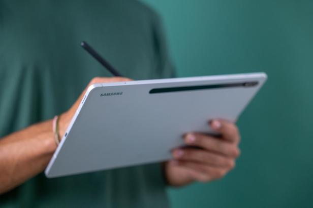 Eine Person hält ein silbernes Samsung-Tablet und nutzt einen Eingabestift vor einem grünen Hintergrund.