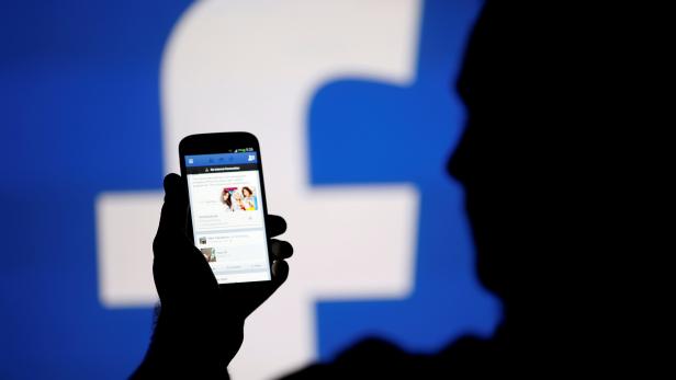 A man is silhouetted against a video screen with a Facebook logo as he poses with a smartphone in this photo illustration