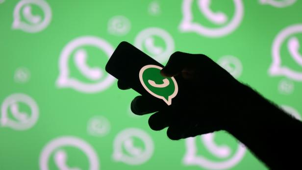 Eine Hand hält ein Smartphone mit dem WhatsApp-Logo vor einem grünen Hintergrund mit WhatsApp-Symbolen.