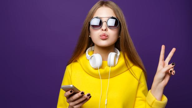 Portrait young girl teenager with earphones and phone, in a yellow sweater, isolate on a violet background.