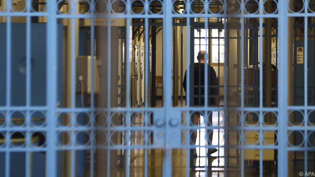 Eine Person steht hinter blauen Gitterstäben in einem Flur mit Zellentüren und Fenstern.