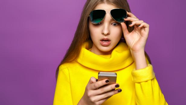 Portrait surprised young girl from message on smartphone in brightly yellow sweater, isolate on a violet background