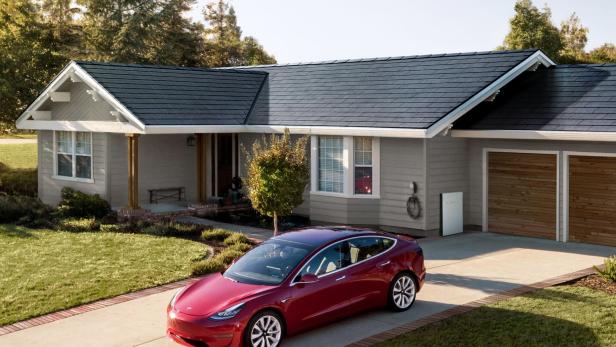 Ein rotes Tesla Model 3 steht vor einem Haus mit Solardach und Tesla Powerwall.