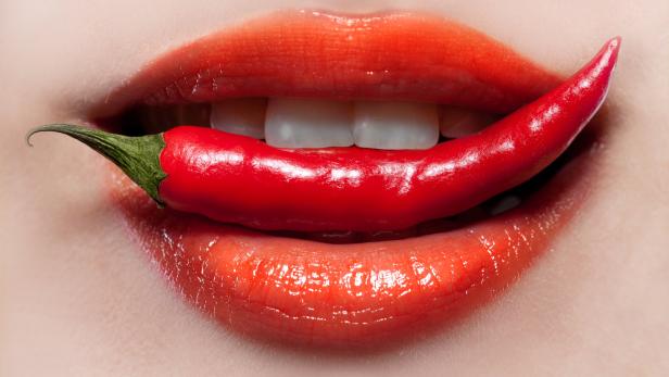 Nahaufnahme von einem Mund mit rotem Lippenstift, der eine rote Chili zwischen den Zähnen hält.
