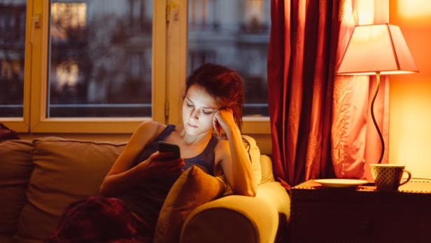 Eine Frau sitzt abends auf einem Sofa und schaut auf ihr Smartphone.