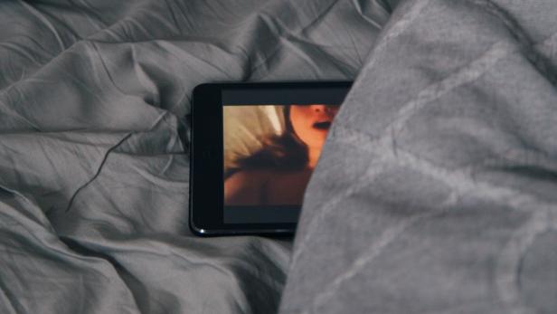 Ein Smartphone, auf dem eine Frau in erotischer Pose zu sehen ist, liegt zwischen grauen Bettlaken