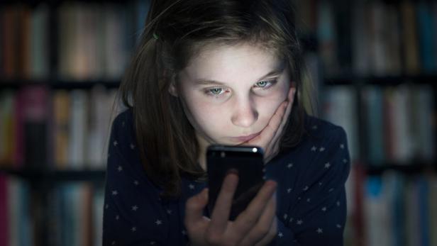Ein Mädchen schaut mit ernstem Blick auf ihr Smartphone, im Hintergrund ein Bücherregal.