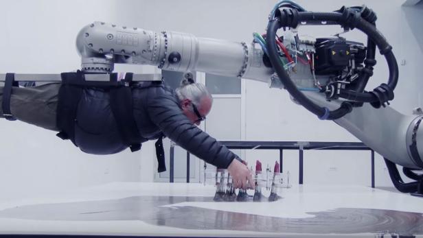 Der Künstler Dragan Ilic hängt an einem Industrie-Roboter-Arm über einer Leinwand