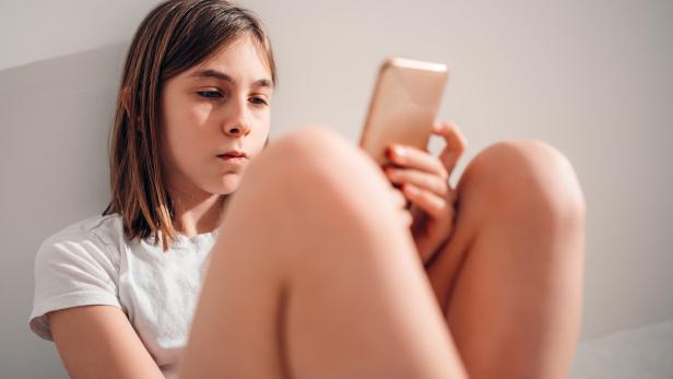 Mädchen mit braunen Haaren sitzt an eine Wand gelehnt und schaut konzentriert auf ein Smartphone in ihrer Hand.