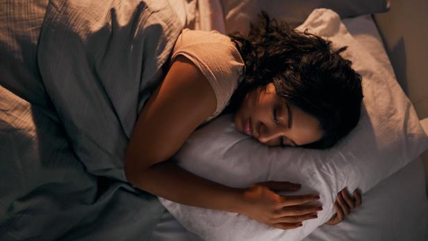 Eine Frau liegt mit geschlossenen Augen bei dämmrigem Licht im Bett.