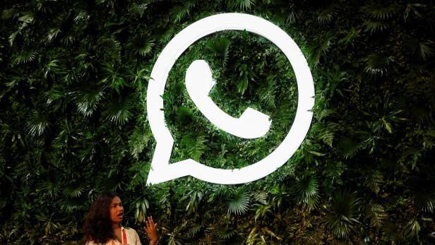 Eine Frau spricht vor einer großen, leuchtenden WhatsApp-Logo-Installation an einer Wand mit vielen grünen Pflanzen.