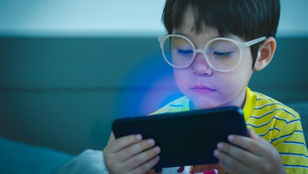 Ein Kind mit Brille sitzt vor einem hell erleuchteten Handy-Display.
