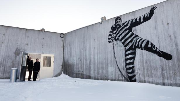 Im Innenhof des Halden-Gefängnisses in Norwegen blicken zwei Aufseherinnen auf ein Wandgemälde, das einen Gefangenen  zeigt, der mit einer Kugel an einer Kette Kugelstoßen übt
