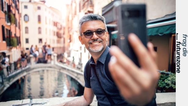 Tourist posiert für Selfie mit Smartphone vor einem Kanal mit Brücke darüber in Venedig