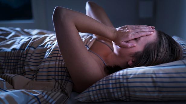Eine Frau liegt im Bett und bedeckt ihr Gesicht mit den Händen, wirkt erschöpft oder gestresst.