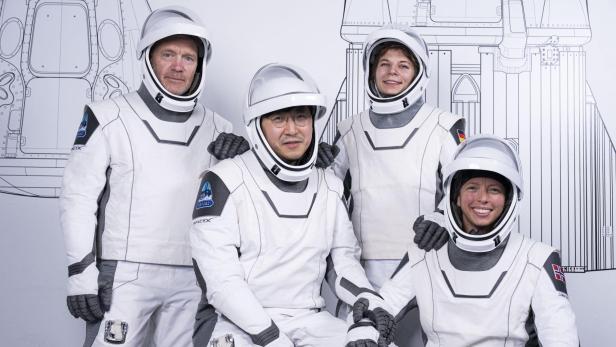 Die Fram2-Crew besteht aus Abenteurer Eric Philips, Crypto-Milliardär Chun Wang, Elektroingenieurin Rabea Rogge und Filmemacherin Jannicke Mikkelsen (v.l.n.r.). vier Menschen in weißen-grauen-Raumanzügen mit offenem Visier vor einer weißen Wand, auf der eine schematische Zeichnung zu sehen ist