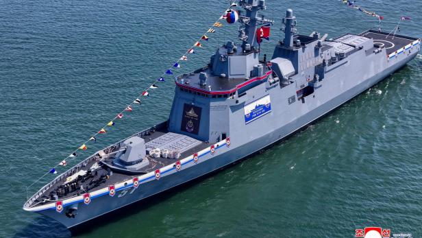Ein großes graues Kriegsschiff mit nordkoreanischer Flagge fährt geschmückt mit bunten Wimpeln auf dem Wasser.