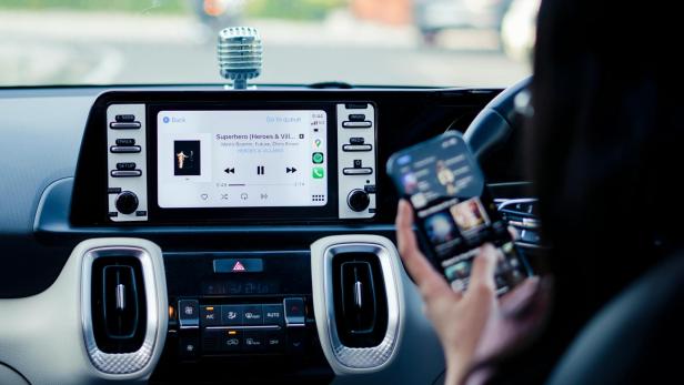 Neben Auto-Bordcomputern sind auch Smart-TVs und Lautsprecher von der Sicherheitslücke betroffen (Symbolbild). jemand hält ein iPhone vor einem Auto-Bordcomputer-Bildschirm