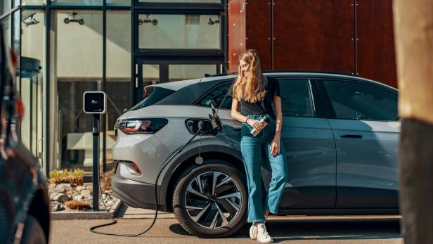 Eine Frau steht lachend vor ihrem Auto, das gerade an einer E-Ladestation aufgeladen wird.
