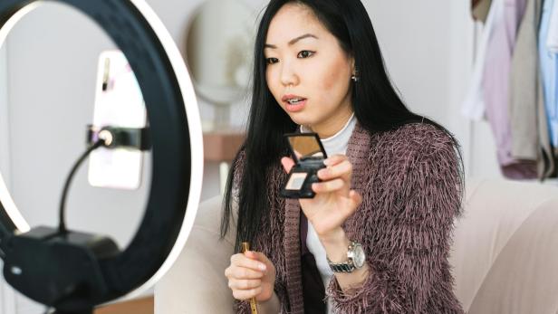 Jugendliche Content Creators unter 16 können nach den neuen Regeln keine Livestreams mehr starten (Symbolbild). eine junge Frau hält eine Make-Up-Schatulle in eine Kamera mit Ringlicht