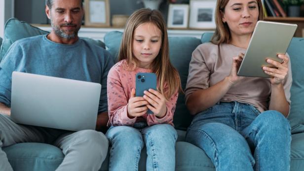 Eine Familie sitzt zusammen auf einem Sofa und nutzt verschiedene digitale Geräte.
